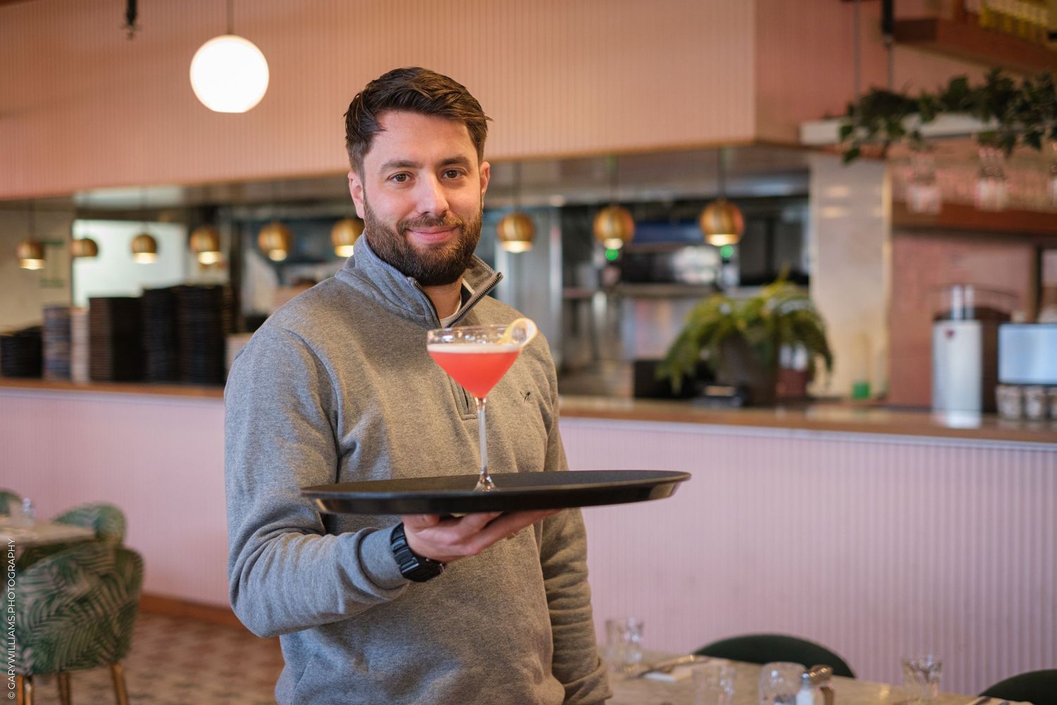 A server carries a colorful cocktail on a tray in a restaurant with pink walls.