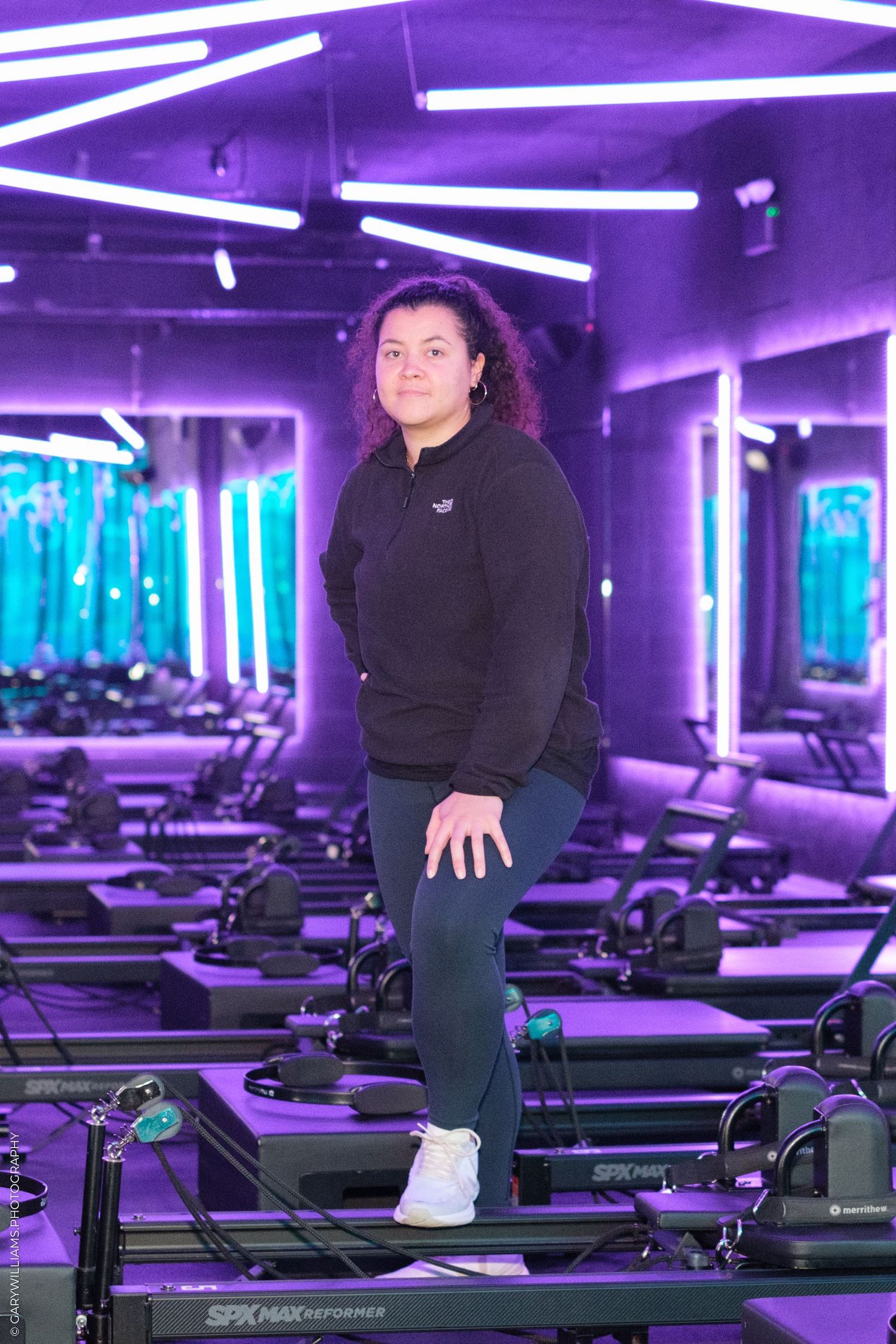 Fitness instructor standing on exercise equipment in a purple-lit gym studio.