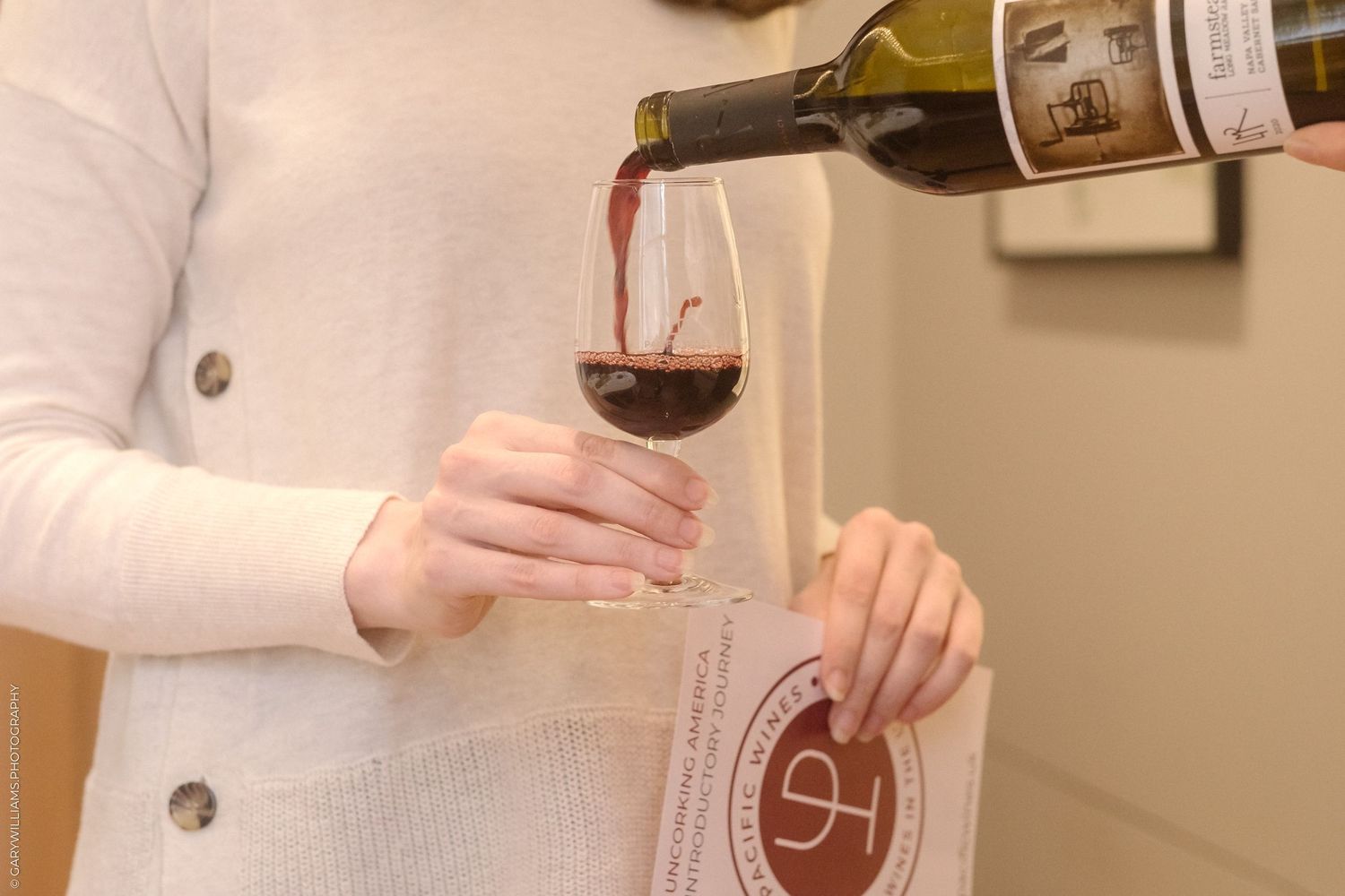 Hands pouring red wine into a glass against neutral background.