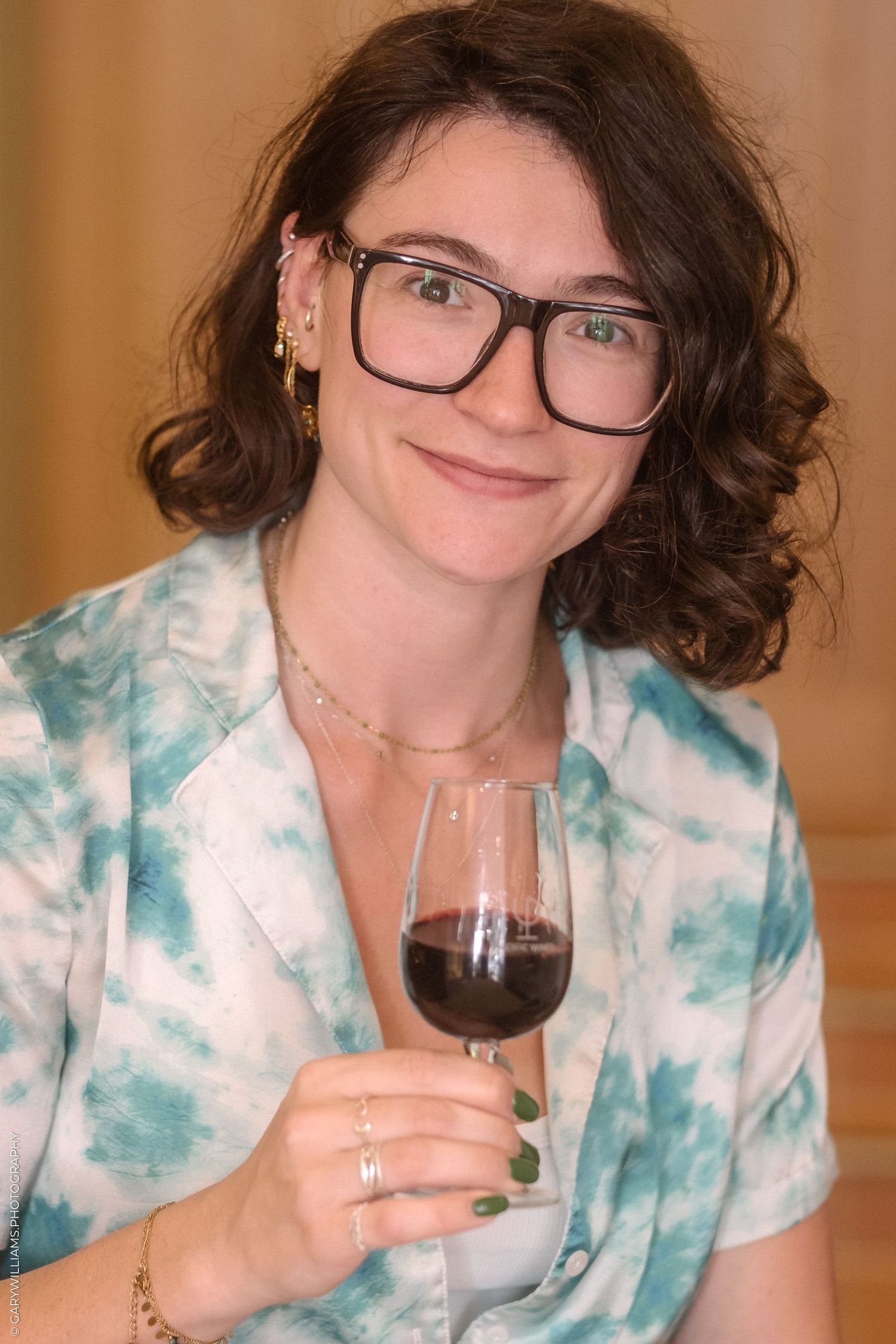 Person in tie-dye shirt holding glass of red wine against warm background.