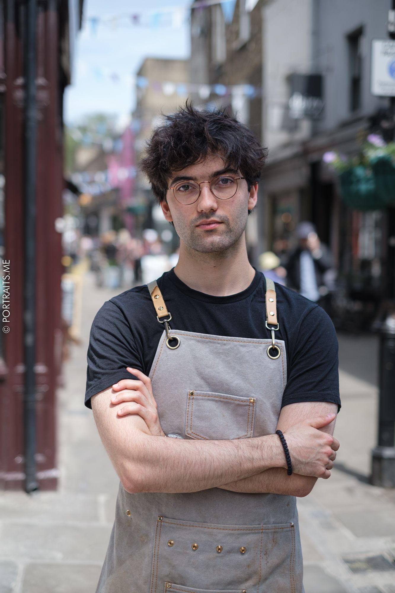 Worker in apron and black shirt poses on city street outside shop.