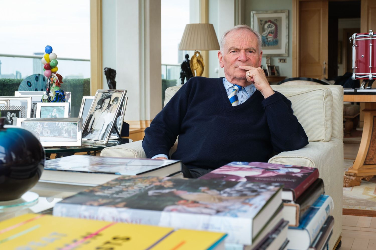 Person in navy sweater sits thoughtfully at desk covered with books.