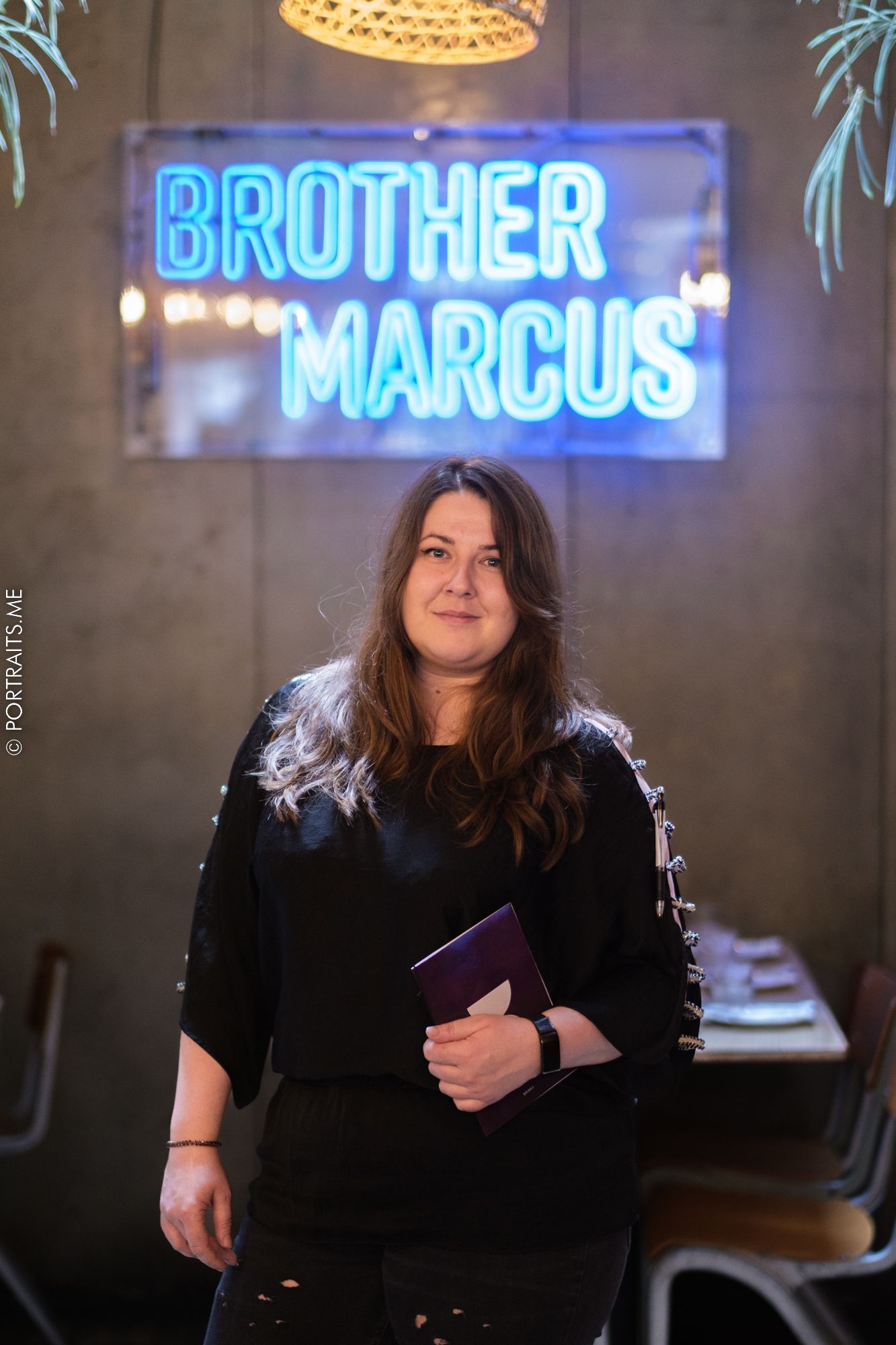 Woman standing beneath bright blue neon sign reading 'Brother Marcus' in restaurant setting.