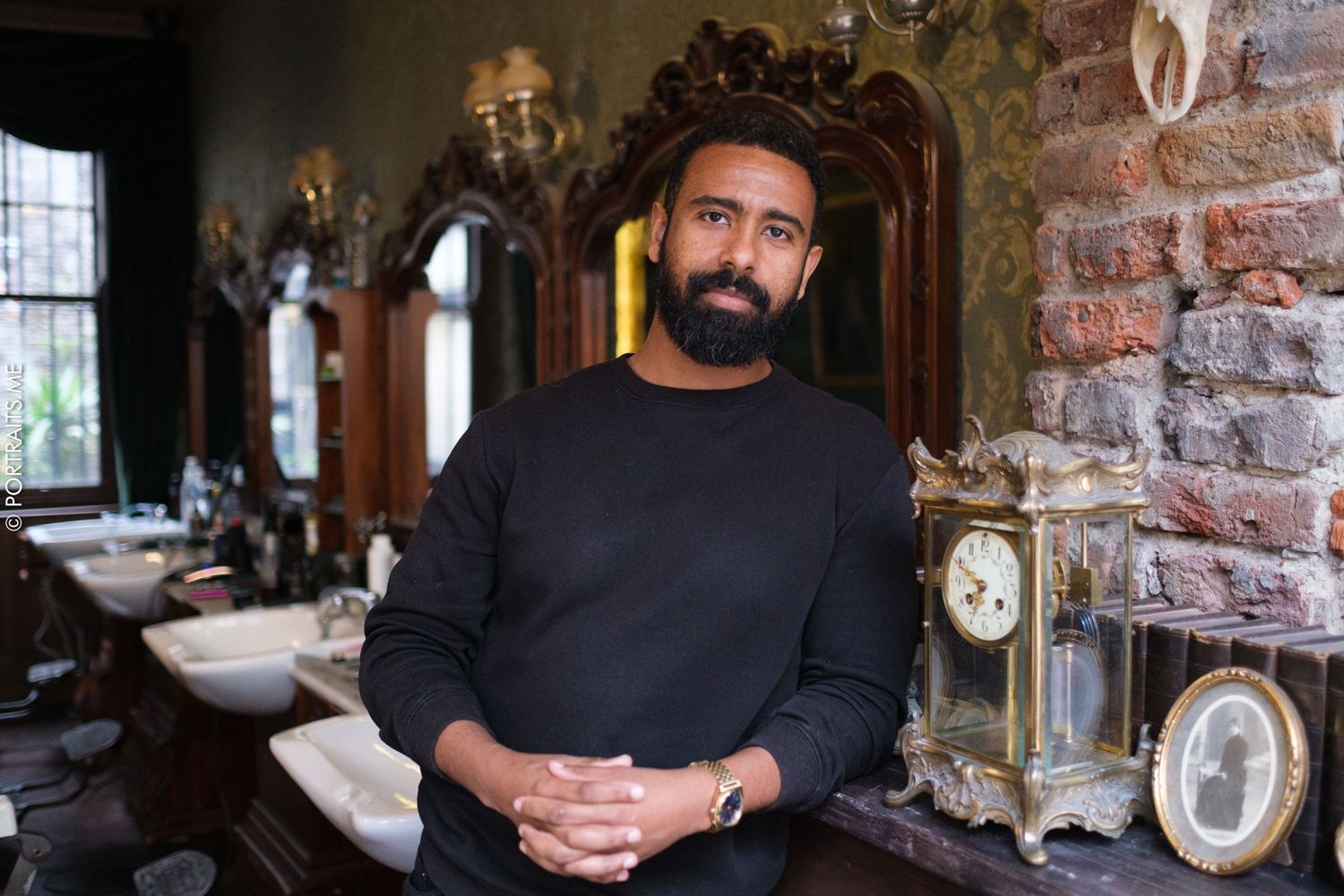 Person in black sweater standing in vintage-styled room with antique decor and exposed brick wall.