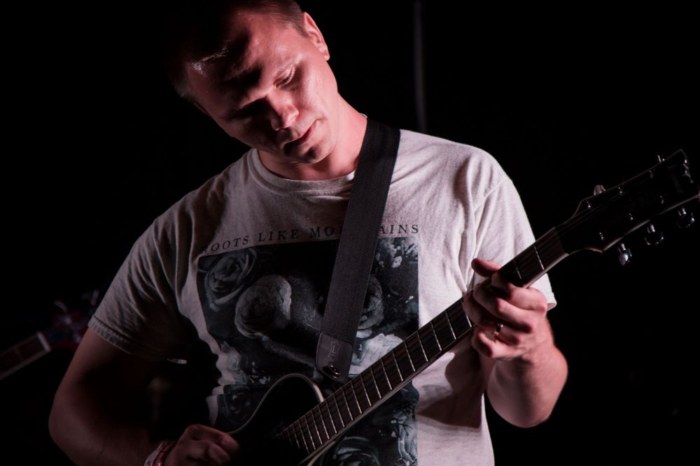 Guitarist performing on stage wearing white t-shirt in dark lighting conditions.