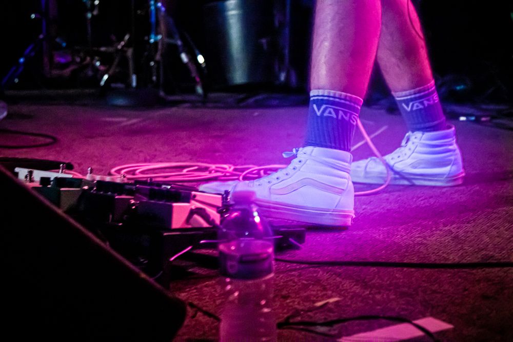 White sneakers and socks illuminated by purple stage lighting near effects pedals.