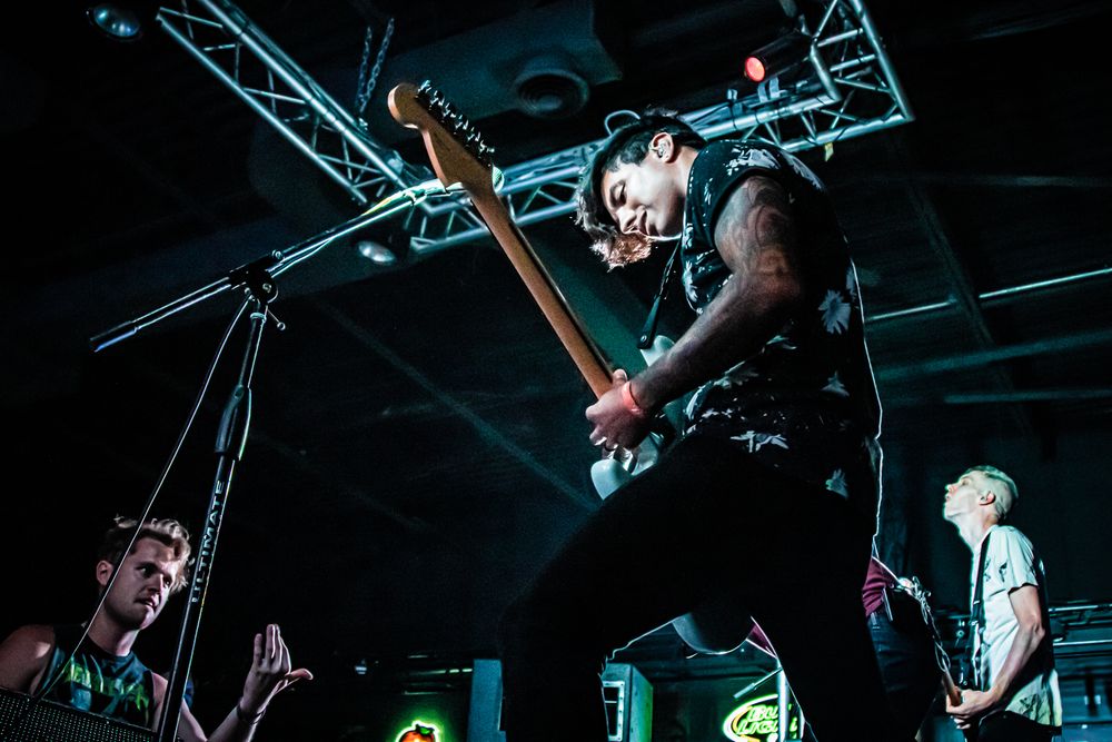 Band members performing energetically on stage with dramatic lighting and truss rigging.