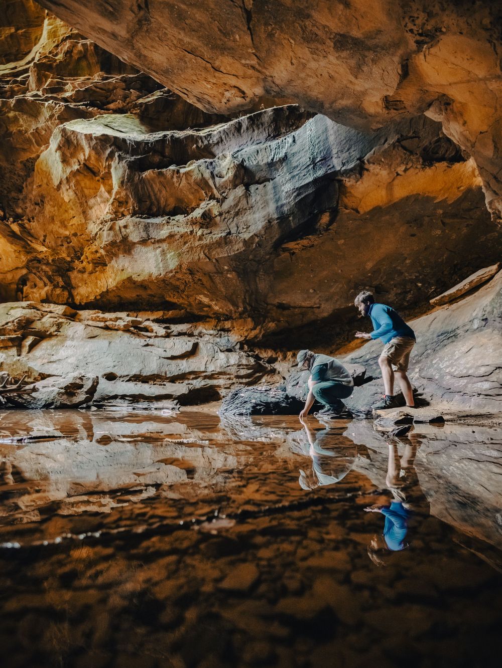 Hikers examining reflective pool inside dramatic cave formation.