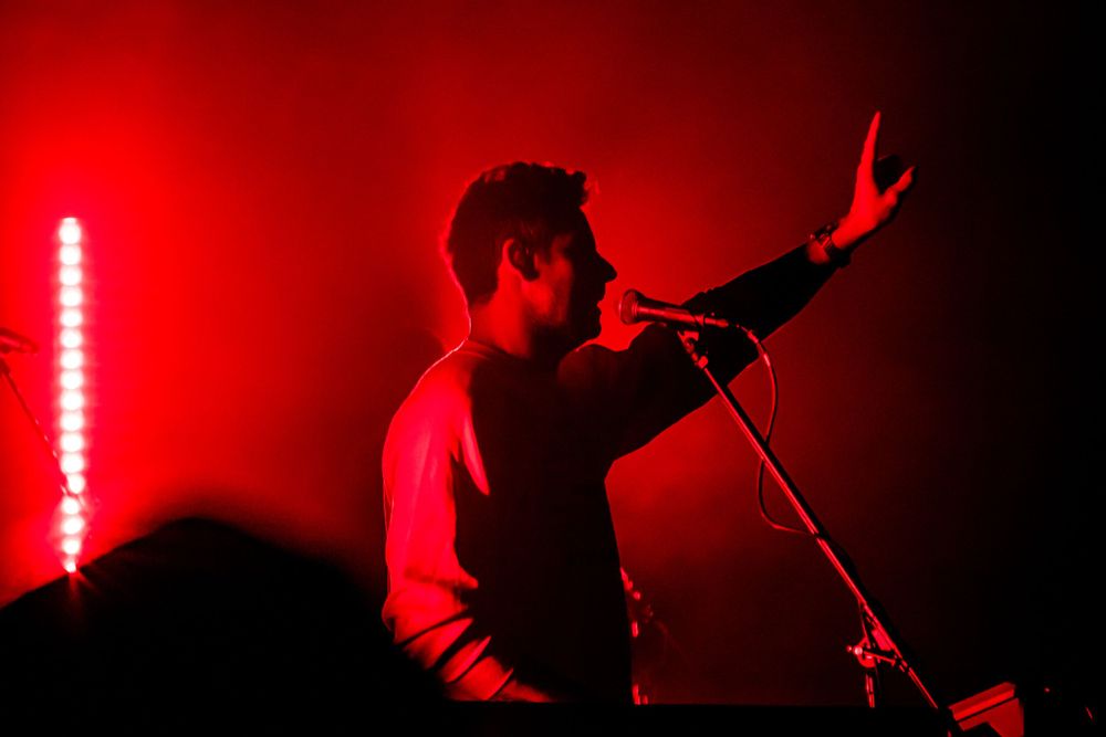 Silhouetted performer singing with raised arm against intense red stage lighting.
