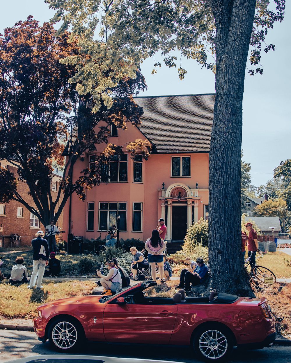 People gathered around red convertible car outside pink building on sunny day.