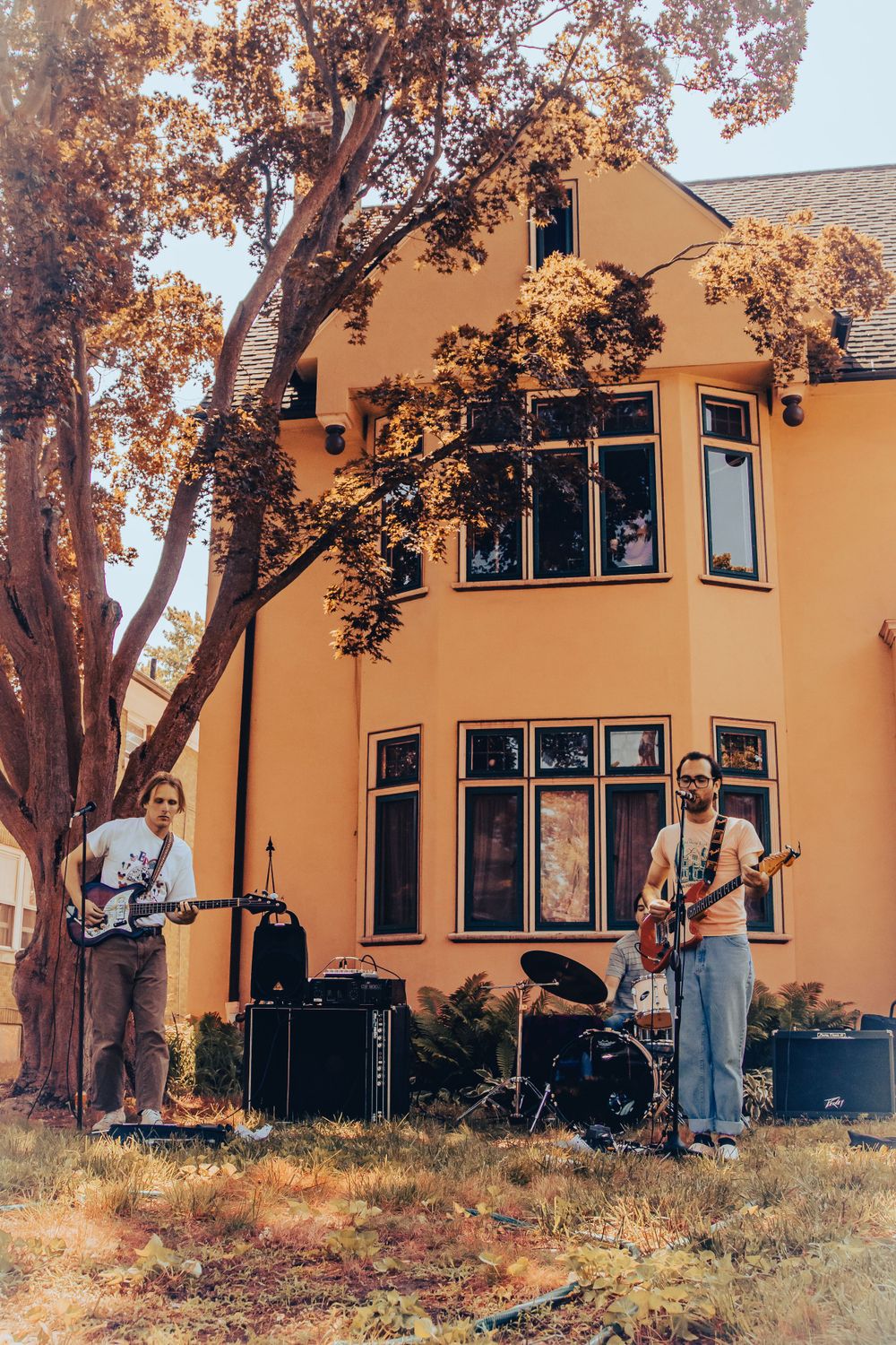 Musicians perform with guitars in front of a peach-colored house with large windows and autumn trees.