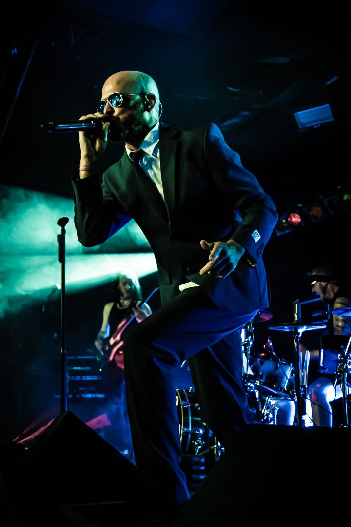 Dramatic concert photo of performer in suit singing on stage with blue and green atmospheric lighting.