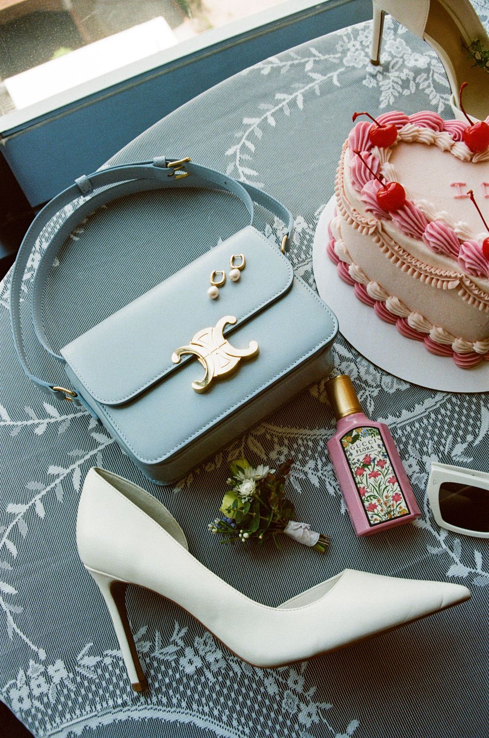 Light blue designer handbag with gold butterfly clasp displayed with white stiletto heel and pink birthday cake on lace tablecloth.