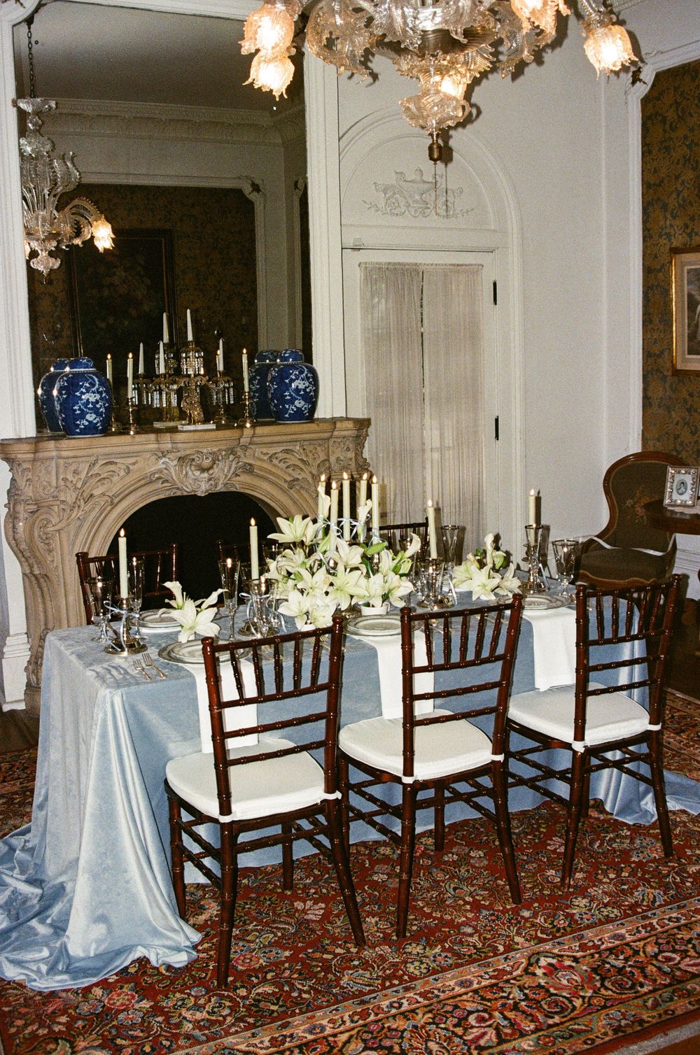 Elegant dining room setup with ornate chandeliers, blue tablecloths, and dark wooden chairs on a patterned oriental rug.
