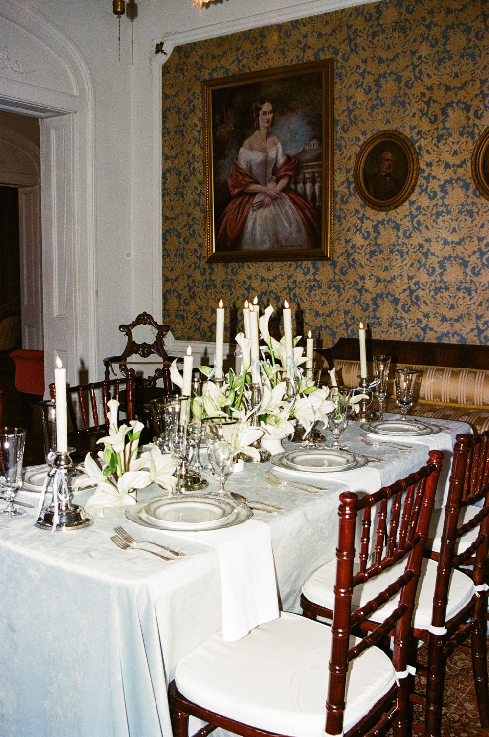 Elegant dining table set with white linens, candles and flowers in a formal room with floral wallpaper and period furniture.