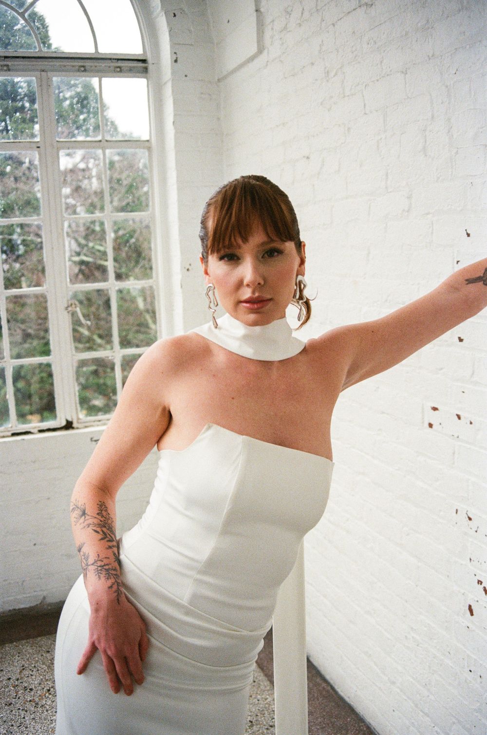 Person wearing a sleek white strapless dress with high neck collar poses near a window in a bright interior space.