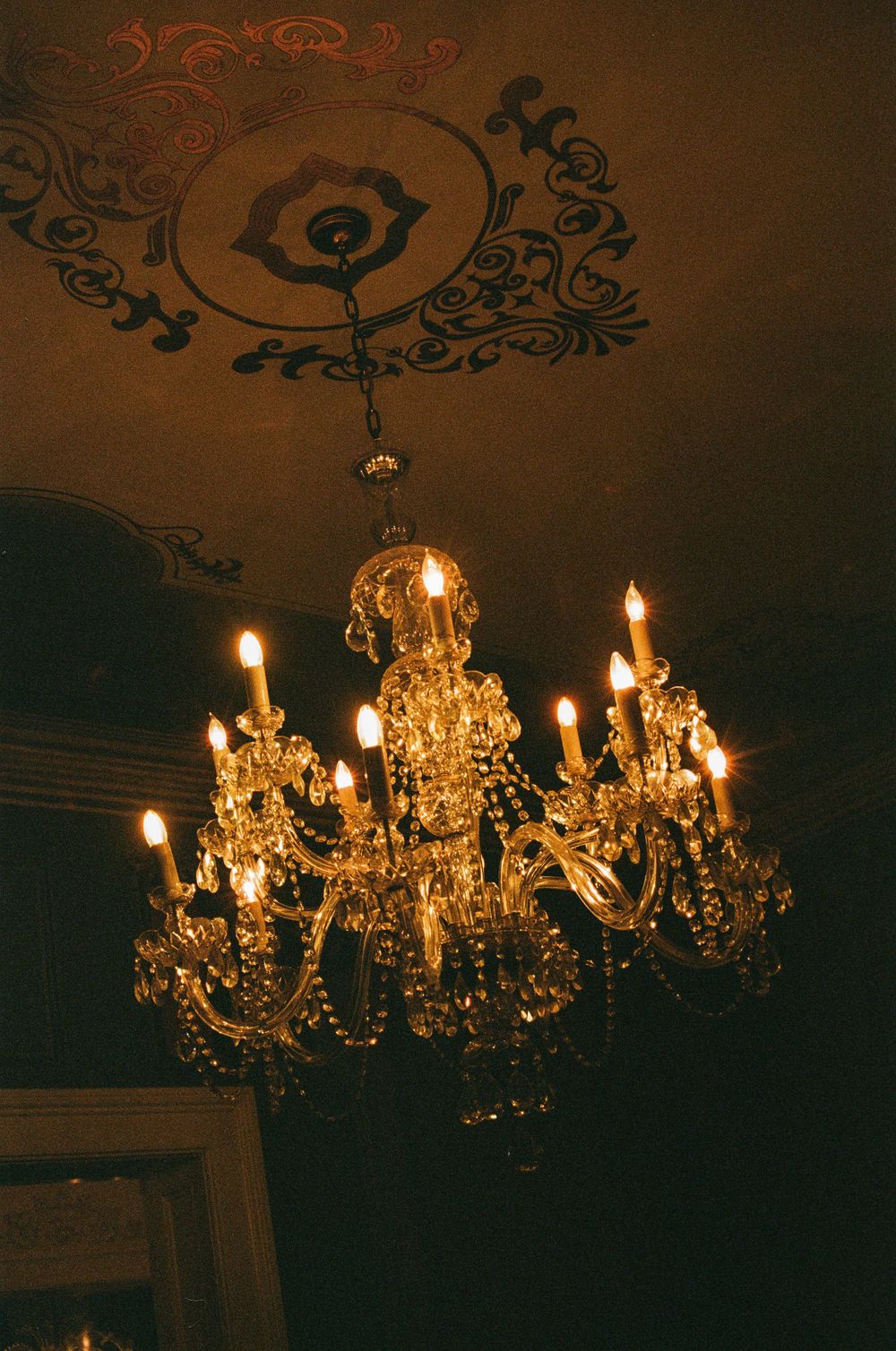 An ornate crystal chandelier with lit candles hangs beneath a decorative ceiling medallion in warm amber lighting.