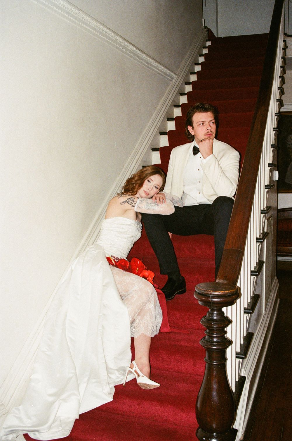 A person in a white dress sits on red-carpeted stairs beside a dark wooden banister in an indoor setting.