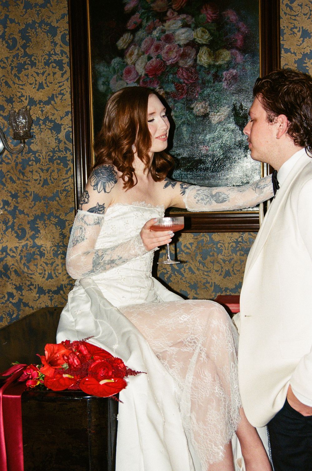 In a vintage room with floral wallpaper and decor, figures in elegant white formal attire share wine near red roses.