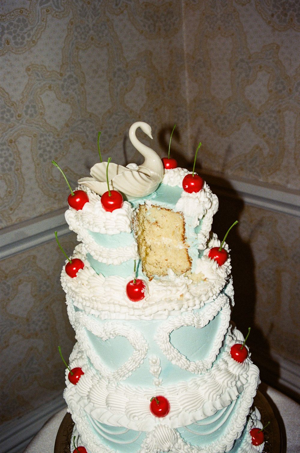 A vintage-style wedding cake with light blue accents, white frosting, and red maraschino cherries topped with a swan decoration.
