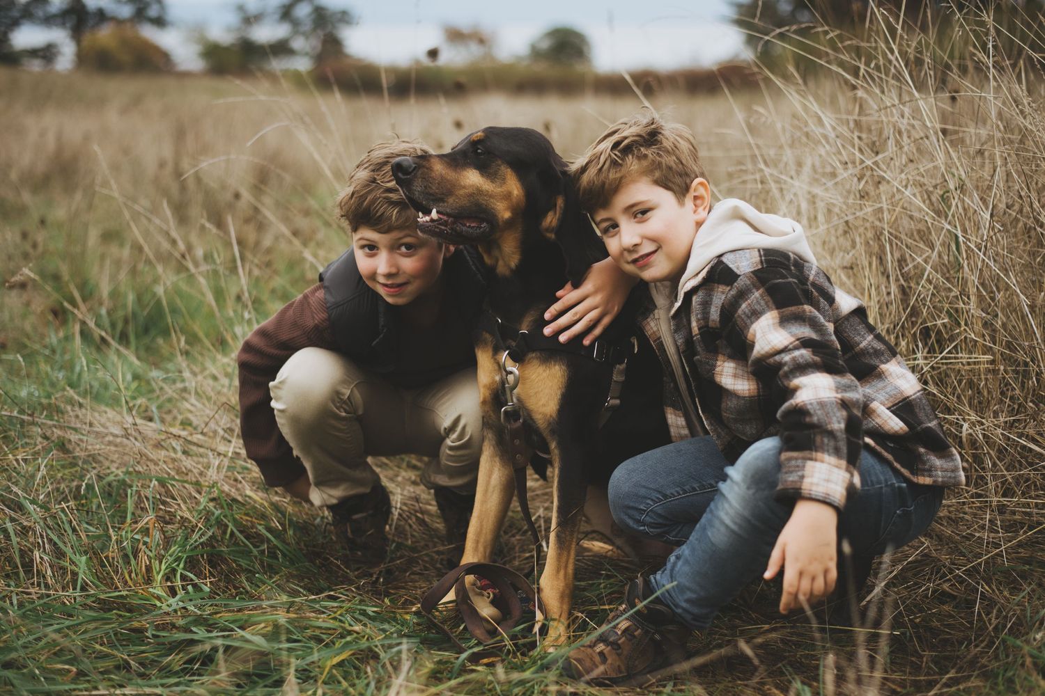 Two young boys crouch down to pose for a photo with their dog in a natural setting.