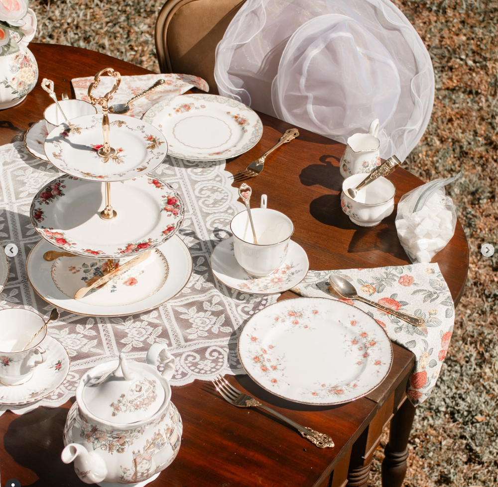 Vintage floral tea set with white porcelain plates and cups arranged on a wooden table with lace tablecloth.