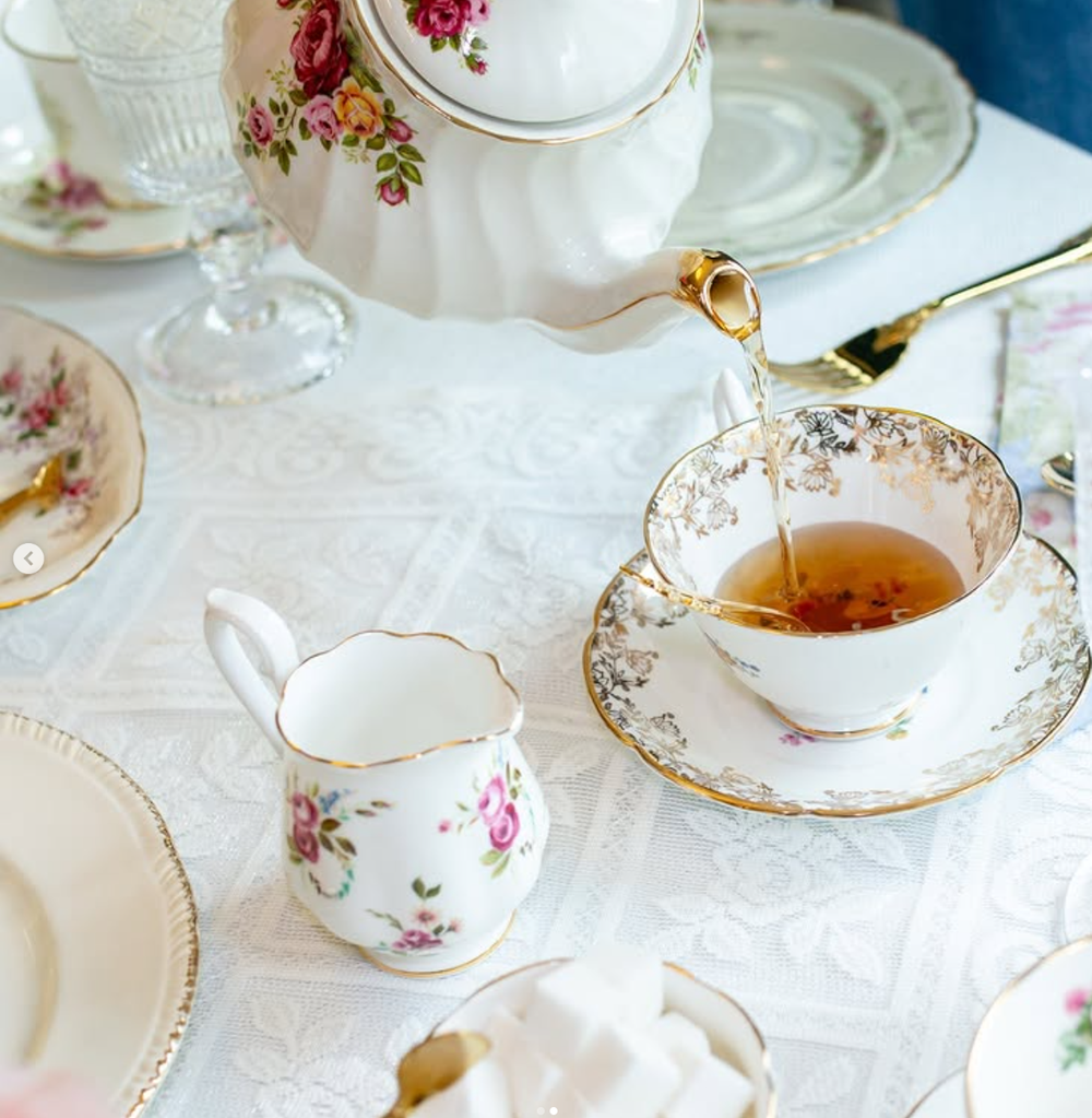 Tea being poured into delicate vintage teacups with floral patterns and gold trim on a white tablecloth.