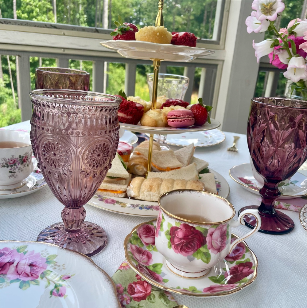 Elegant afternoon tea setup with purple glassware, floral teacup, and sweet treats on tiered serving plates by a window.