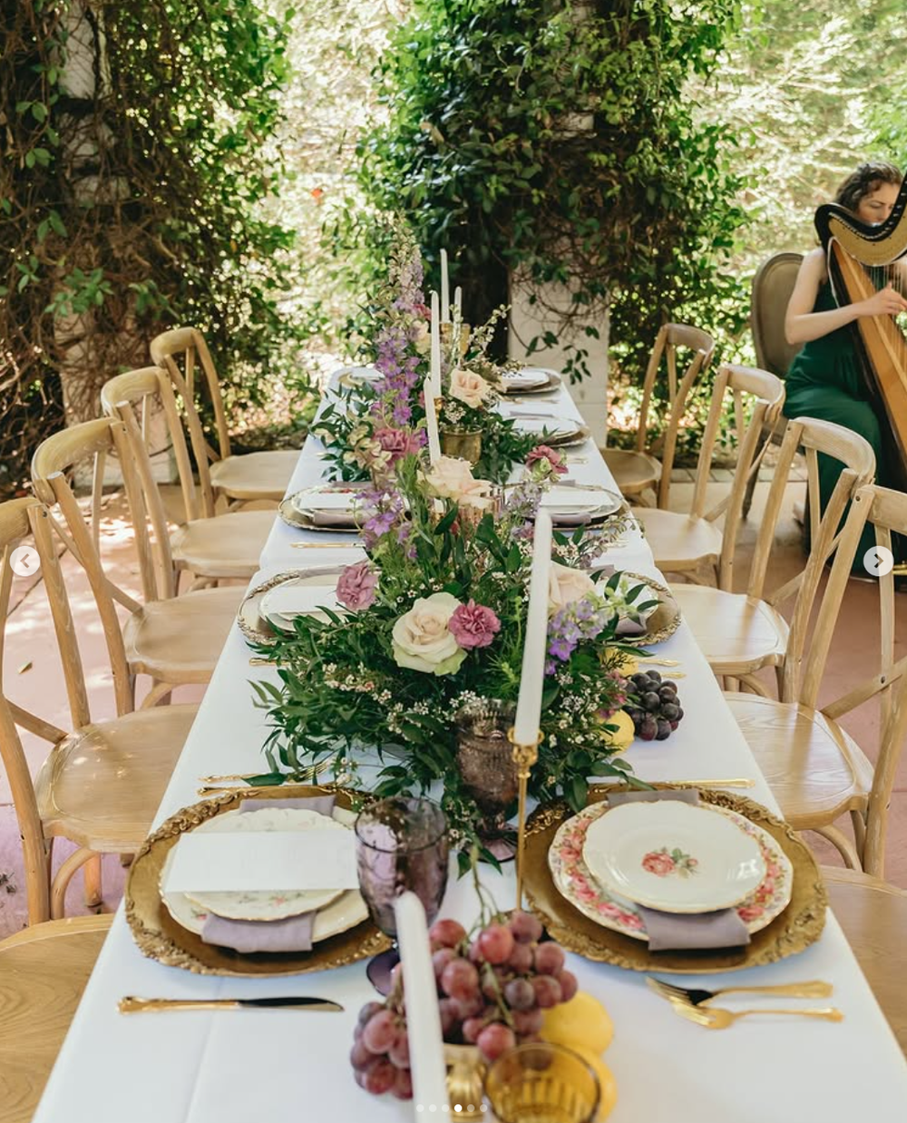 An elegant outdoor table setting with white linens, gold chargers, and purple floral centerpieces under a lush garden canopy.
