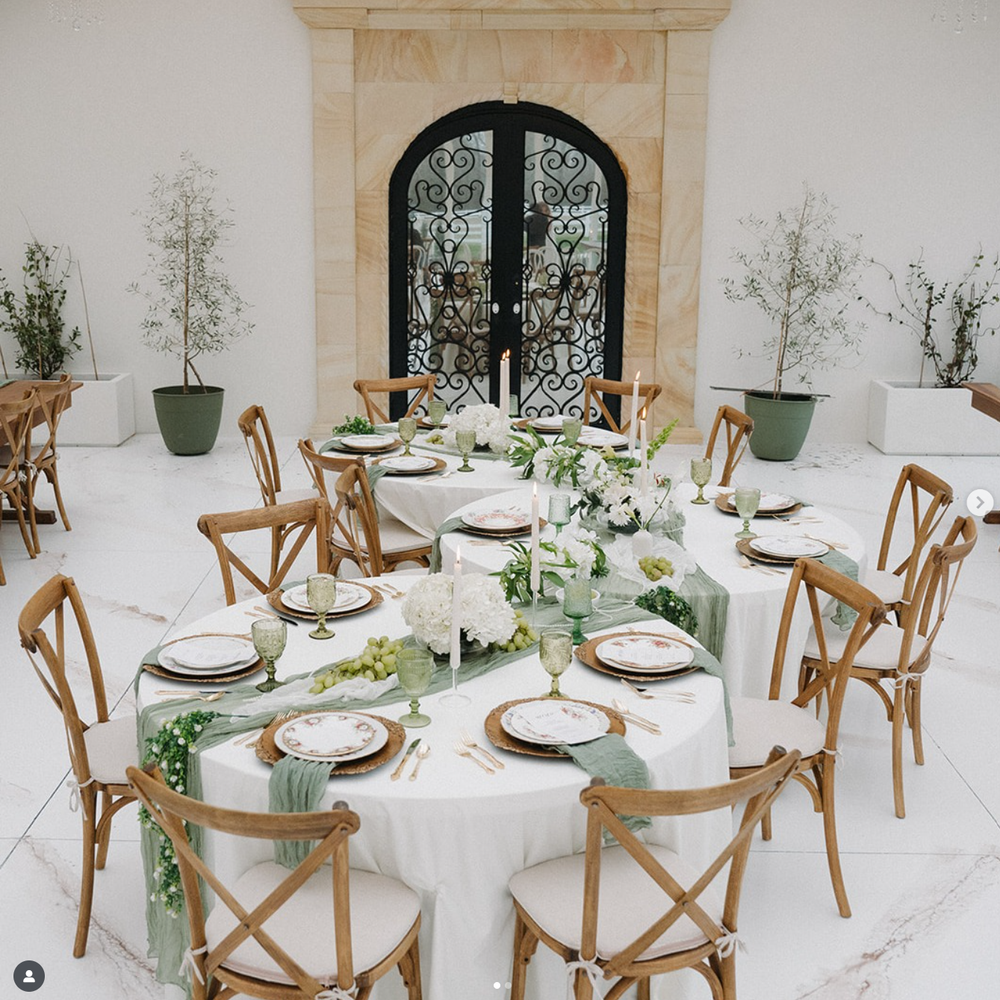 Elegant wedding reception table setup with wooden cross-back chairs and minimalist white table settings with greenery accents.