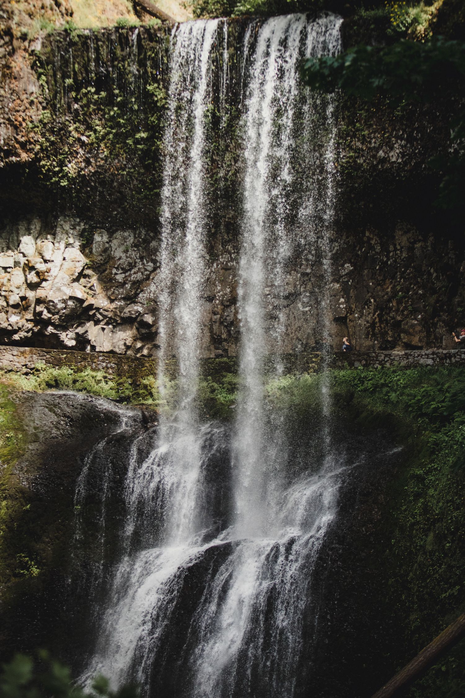 A majestic waterfall cascades down a moss-covered rock face in multiple tiers through lush green surroundings.