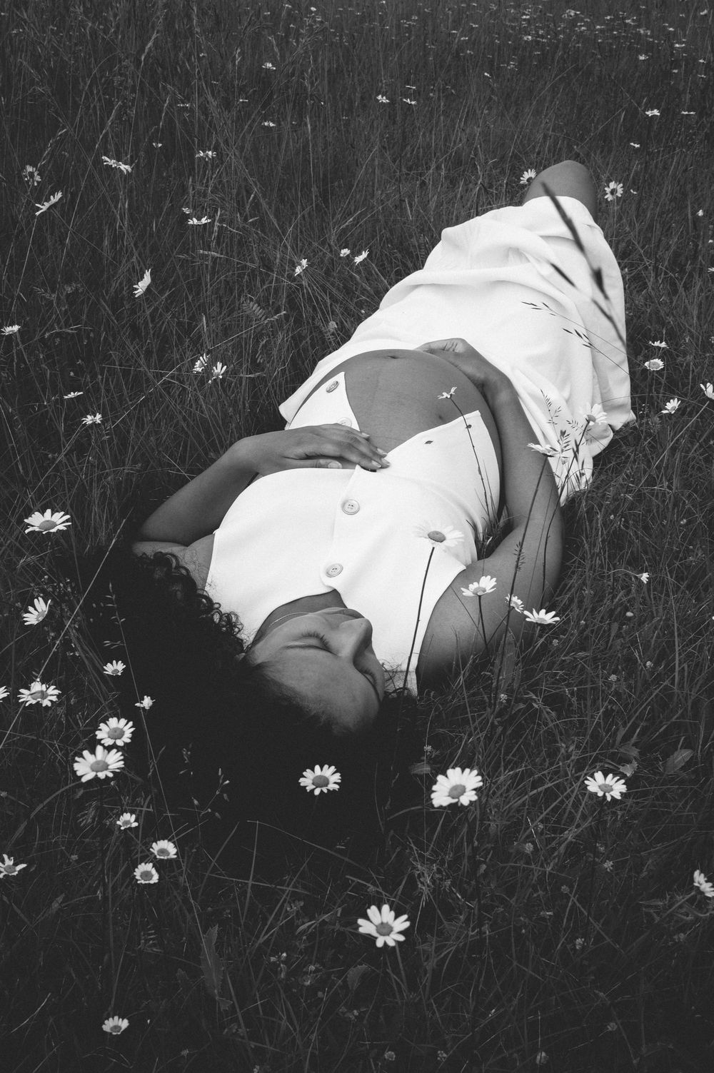 A black and white photograph shows someone lying in a wildflower meadow wearing a light-colored dress among daisies.