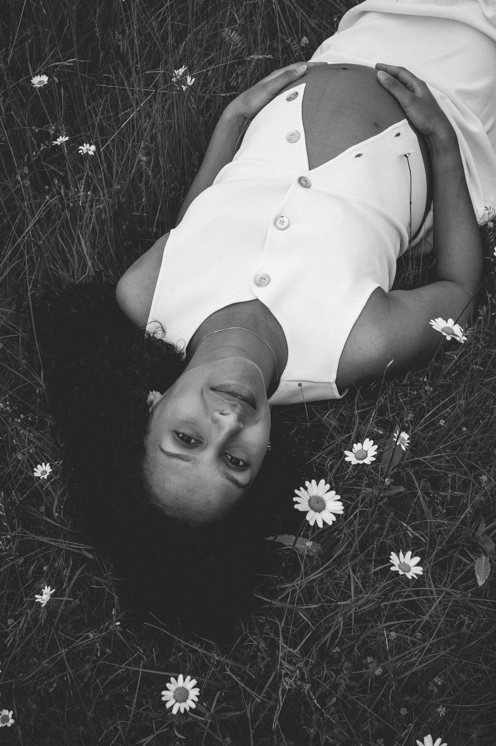Black and white portrait of someone lying back in grass surrounded by delicate white daisies wearing a white blouse.
