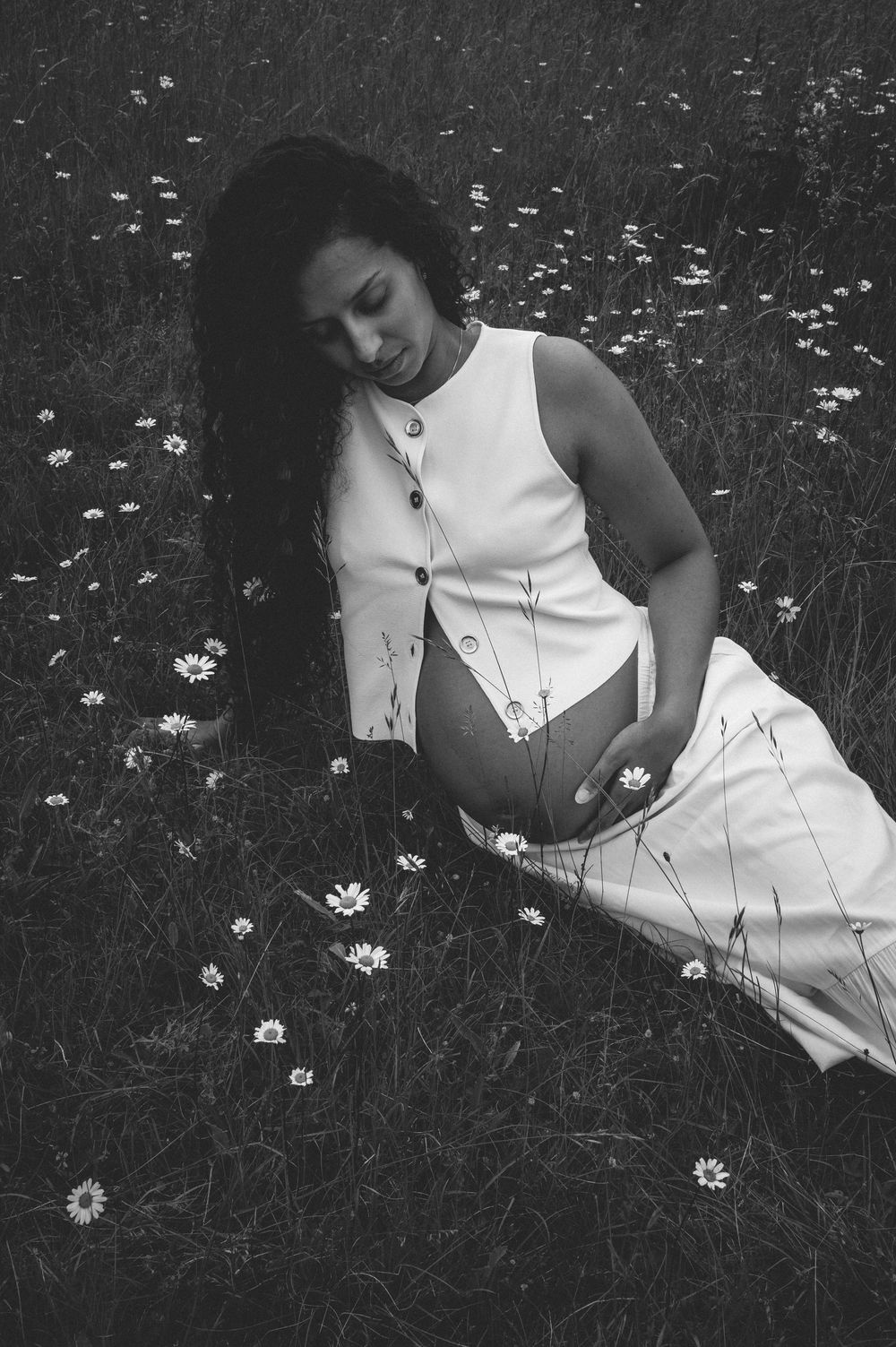Black and white photograph of someone in a white sleeveless dress lying in a field of daisies.