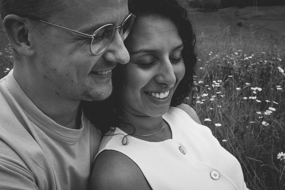Black and white photo shows a couple sitting together and smiling in a field of wildflowers.