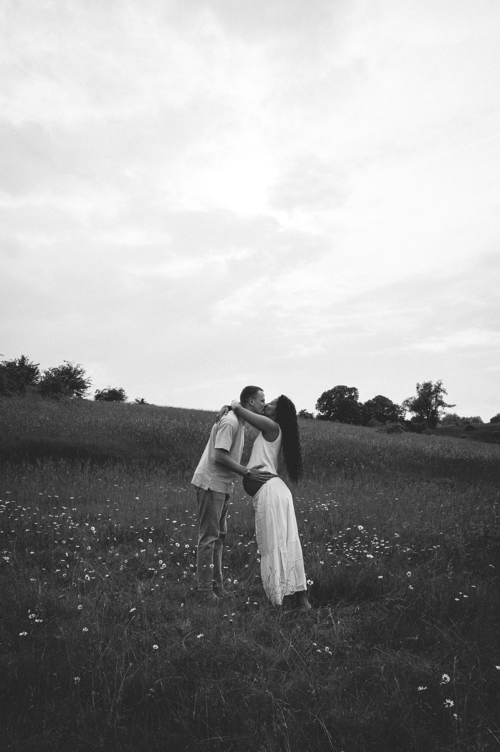A romantic black and white photograph of a couple embracing in a wildflower meadow on a cloudy day.