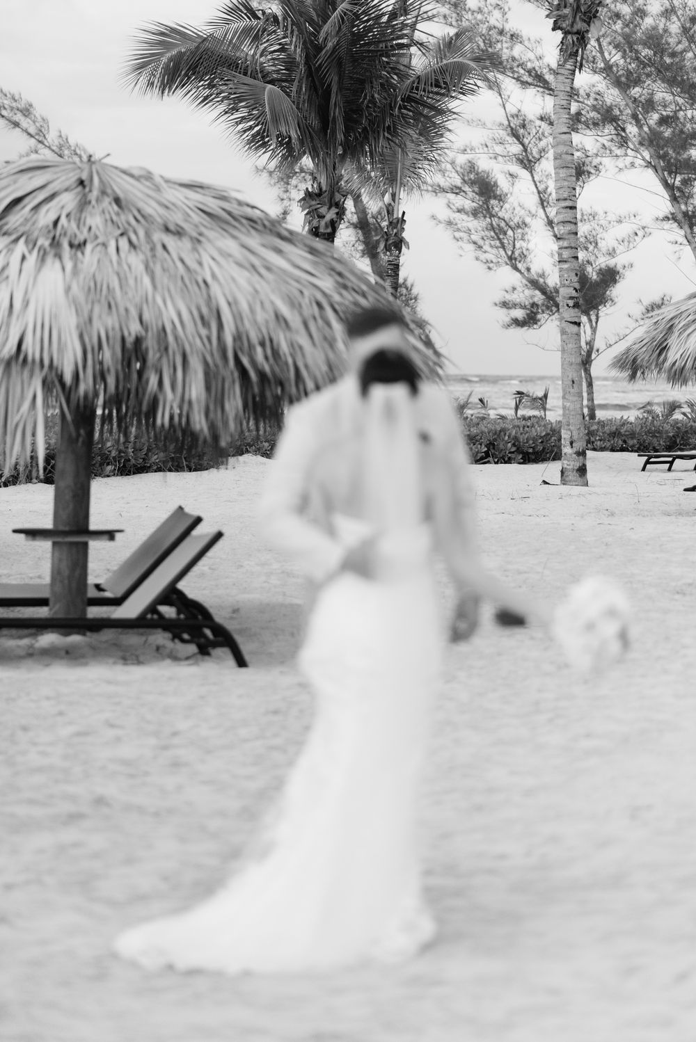 A black and white beach wedding photo with palm trees and thatched umbrellas on a tropical shoreline.