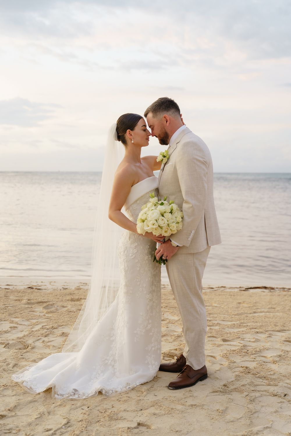 Couple in wedding attire shares a romantic moment on a sandy beach with white bouquet at sunset.
