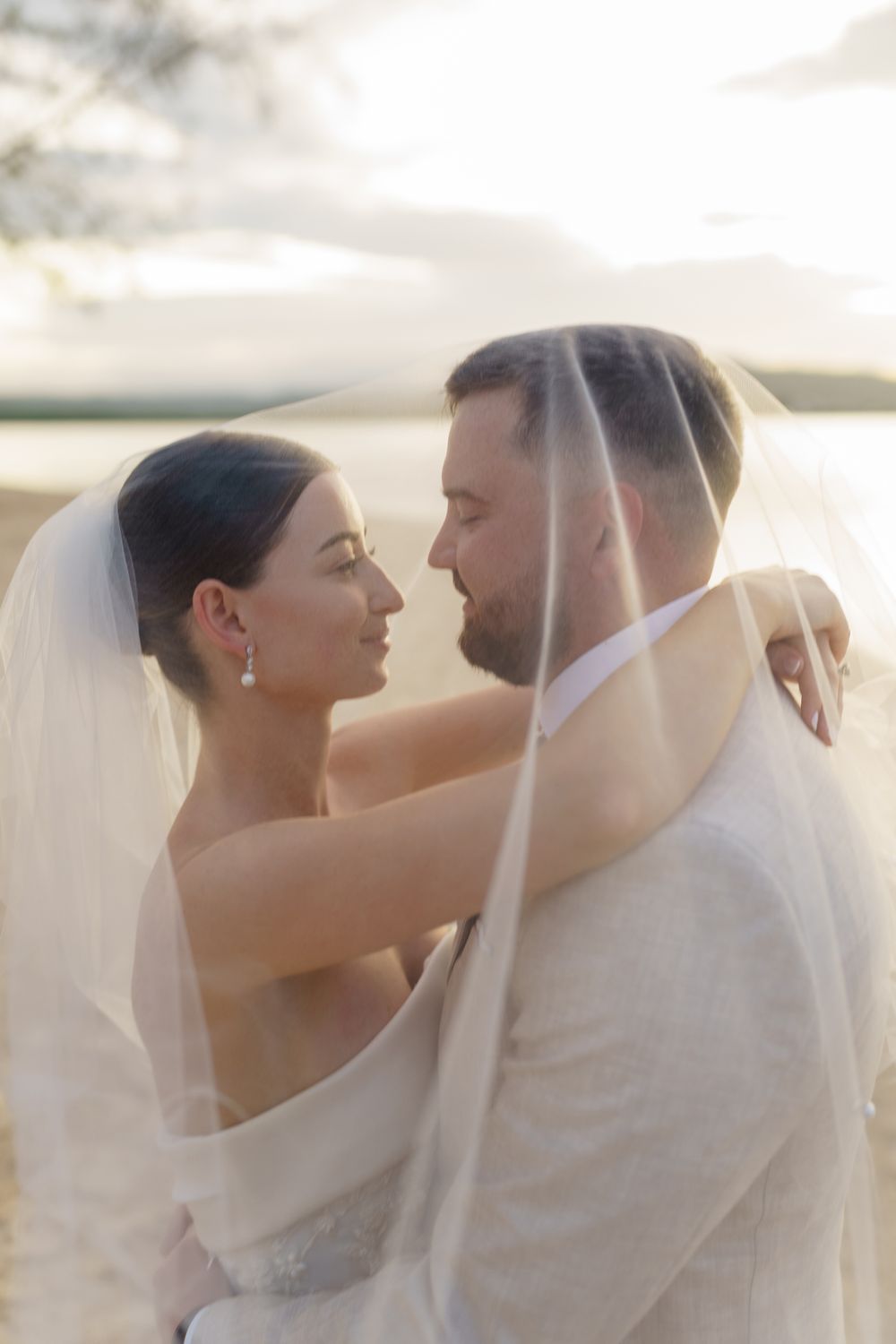 A romantic couple embraces under a flowing veil on a beach during a tender wedding photoshoot.