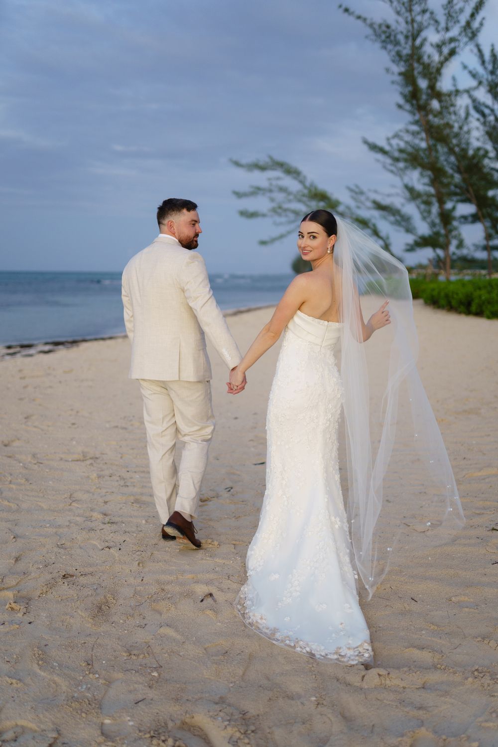 A couple in white wedding attire walks hand in hand on a tropical beach with ocean and trees in background.