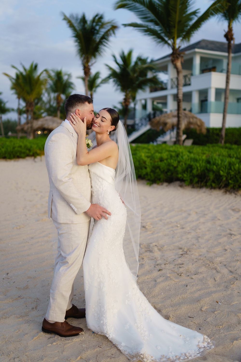 Newlywed couple embraces on tropical beach with palm trees and resort building in background.