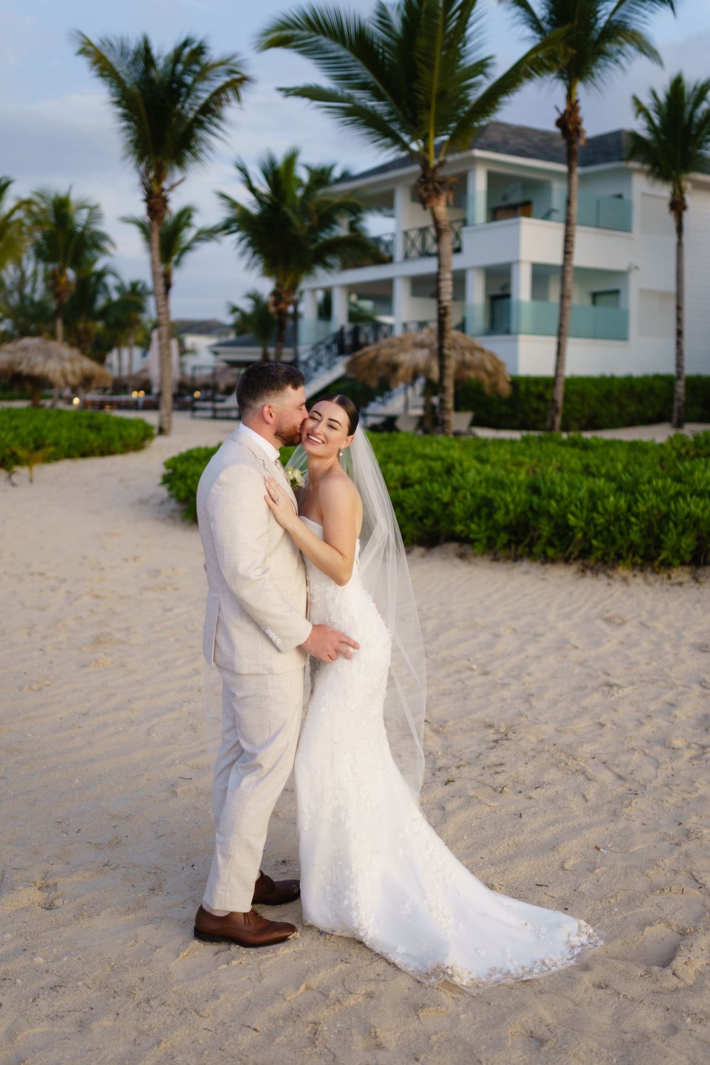 A romantic beach wedding scene with palm trees and a resort building in the background on a tropical sandy shore.