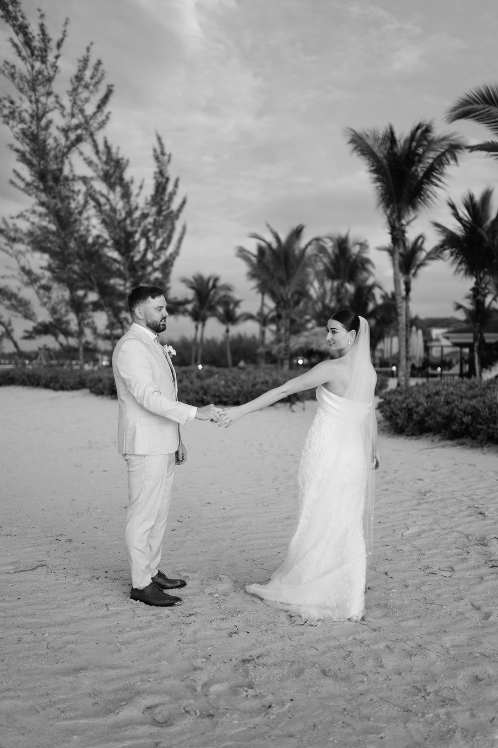 A couple in white attire holds hands on a tropical beach with palm trees and ocean views in black and white photography.