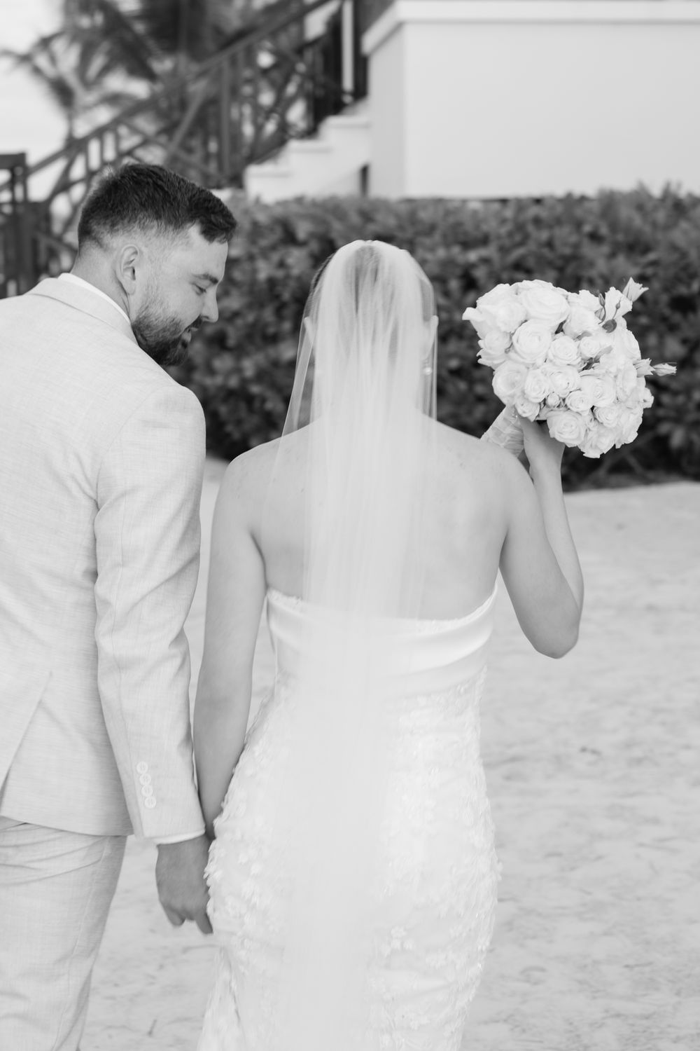 Bride and groom in elegant white attire share romantic moments on a beach in a series of black and white photographs.