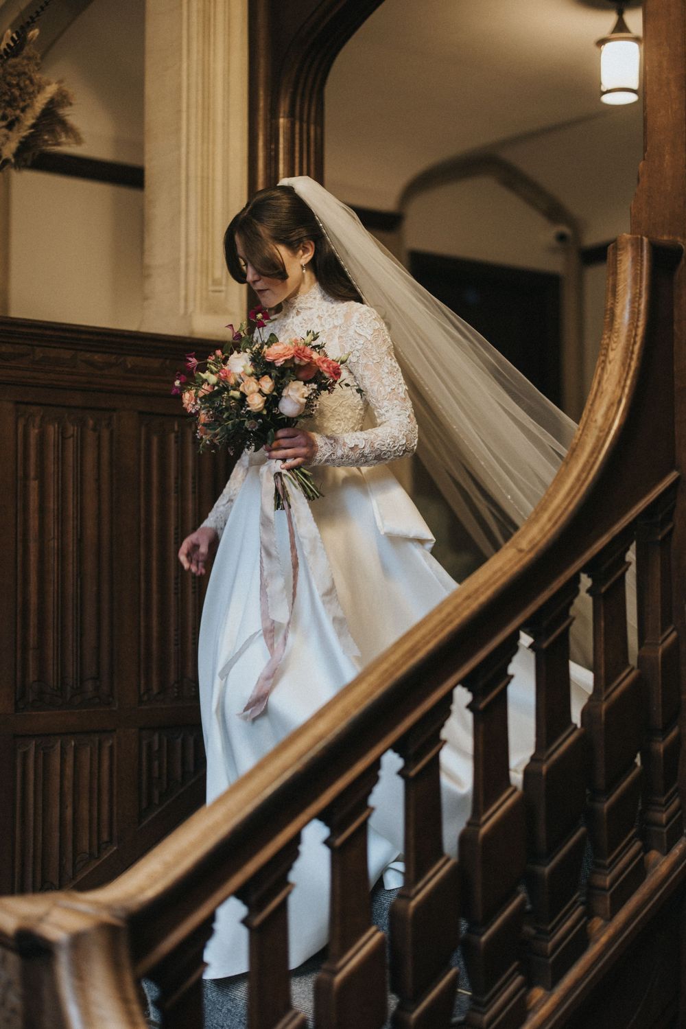 A bride in a white wedding gown descends an elegant wooden staircase while holding a pink and white bouquet.