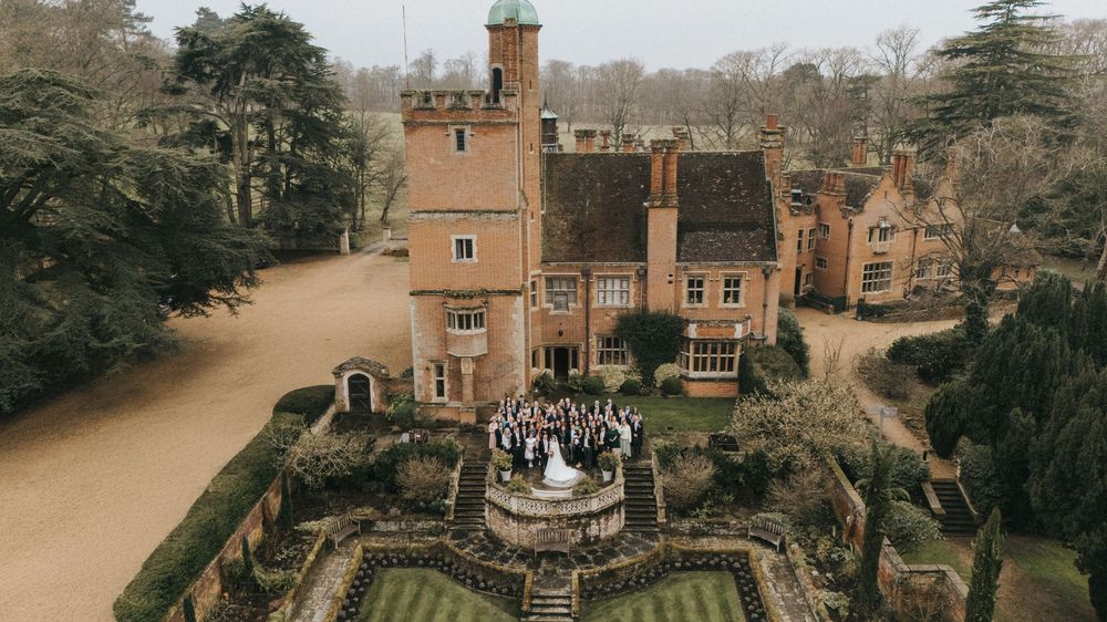 Aerial view of an English manor house with ornate gardens and a gathering of guests on the stone patio.