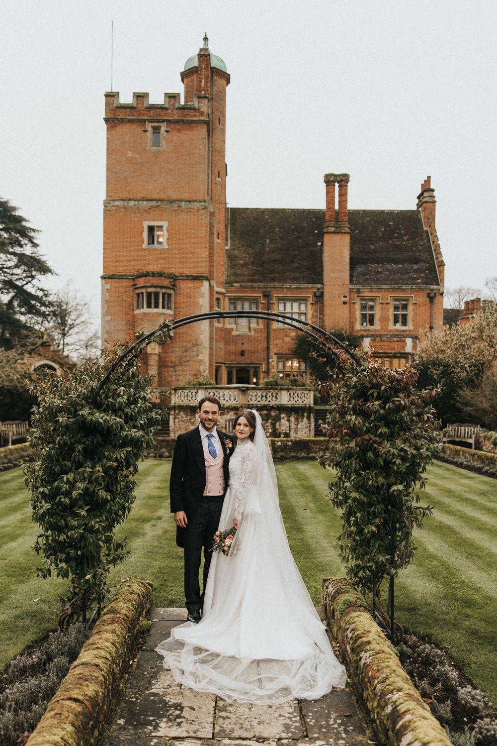 A couple stands on a garden path under an archway at a historic brick manor house with a tower.