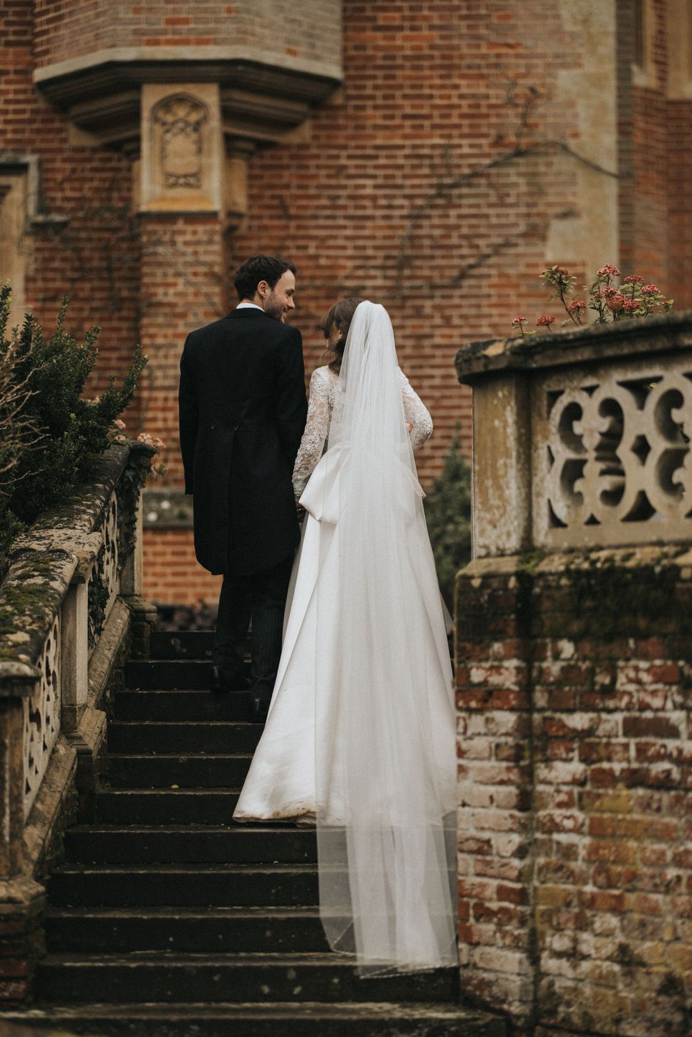 A bride in white gown and groom in black suit stand on vintage brick stairs with ornate stone railings.