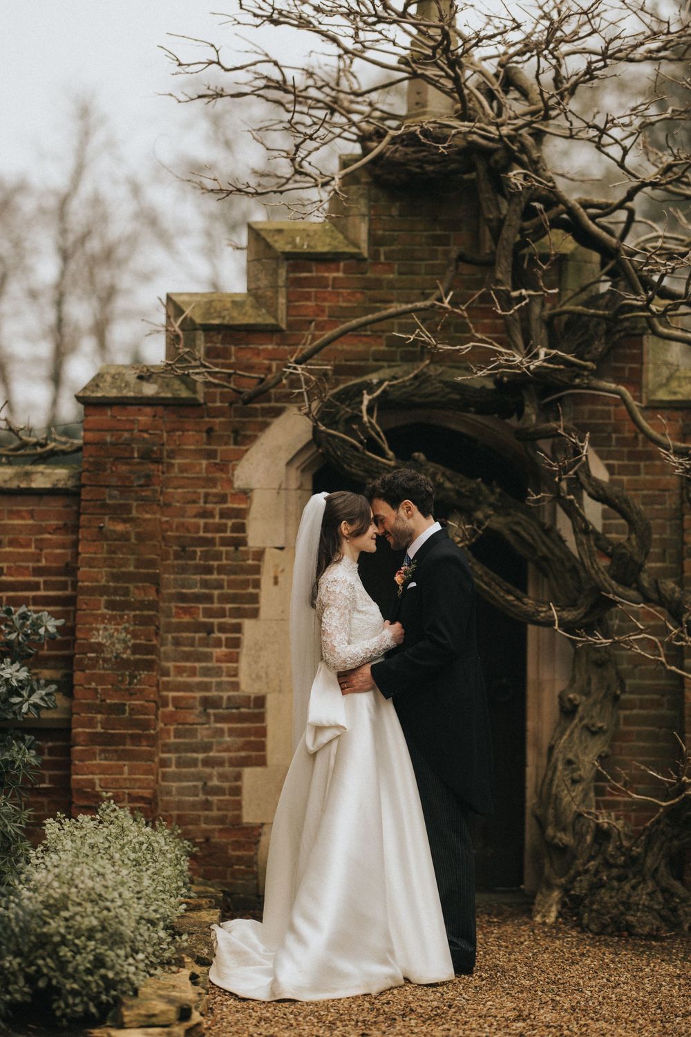An elegant couple shares an intimate moment in front of a rustic brick archway surrounded by bare winter branches.