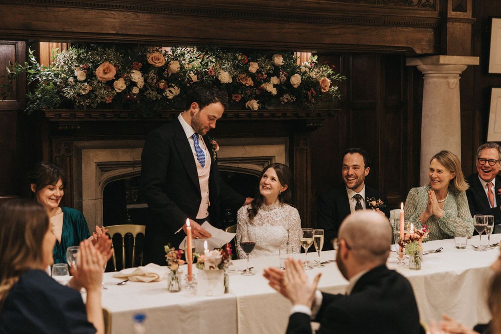 Wedding reception guests applaud during a toast at an elegant venue decorated with peach and white floral arrangements.